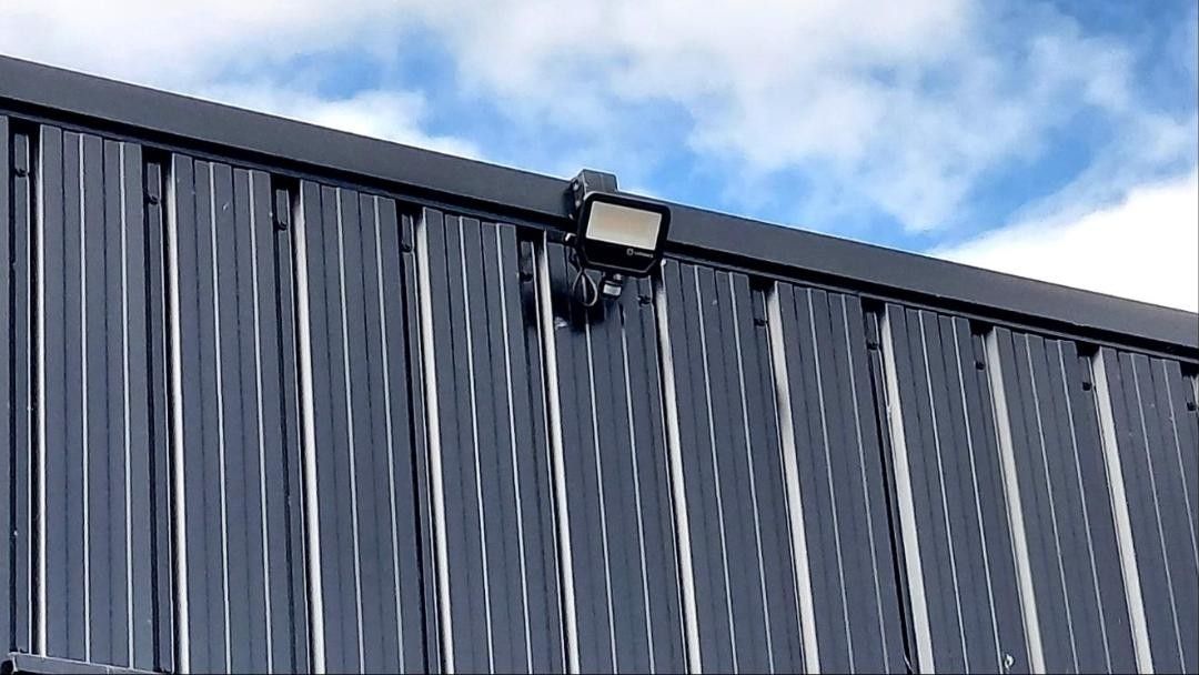 Zwarte LED-schijnwerper met sensor aan de dakrand van een donkere metalen gevel