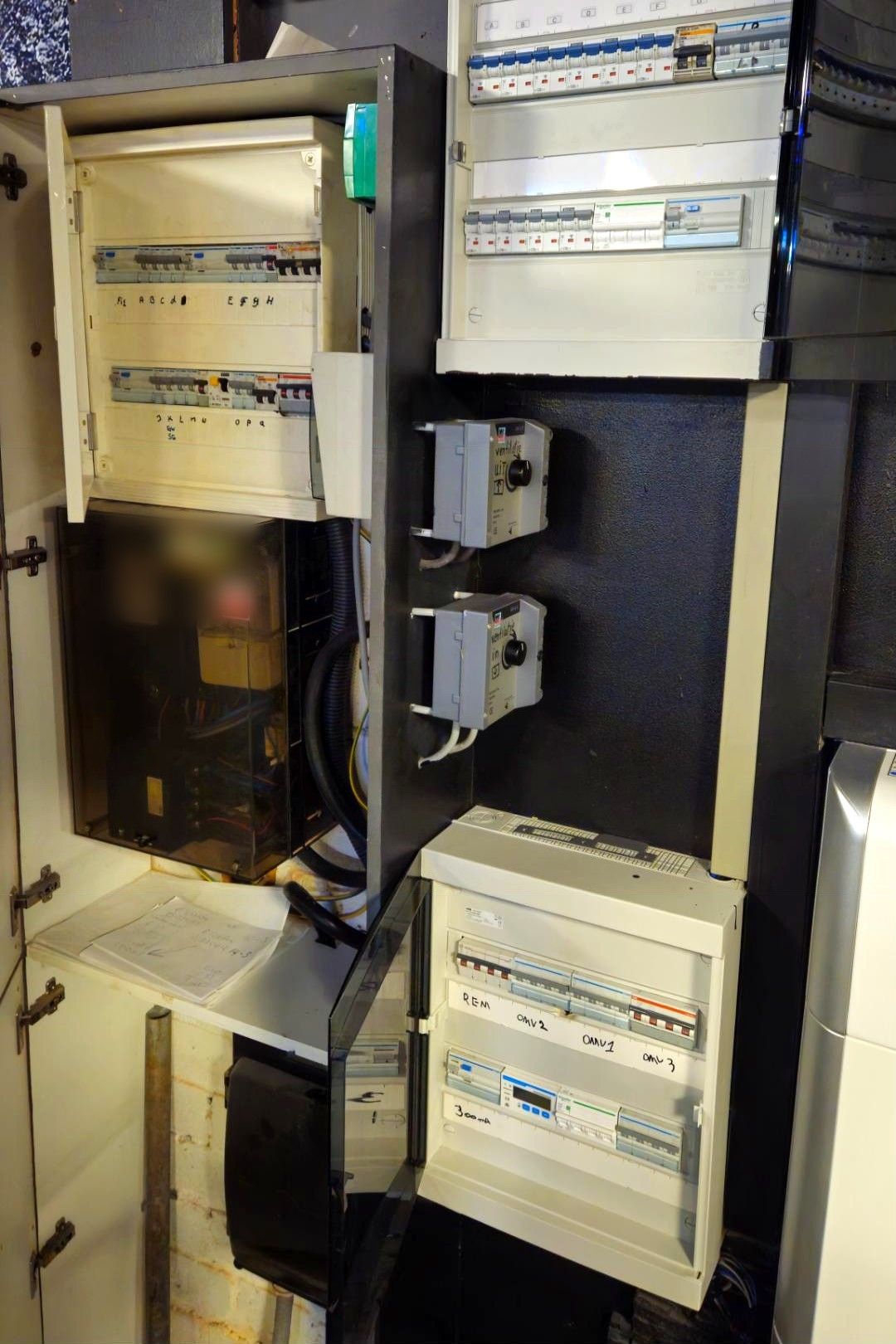 Technische wand met drie oude verdeelborden, een meterkast en twee schakelaars, met handgeschreven letters onder de automaten