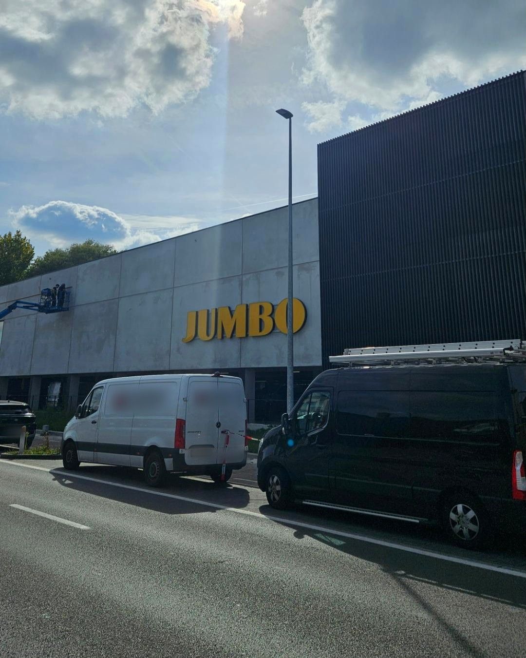 Donkere bestelwagen met ladder op het dak langs de weg, met op de achtergrond de gevel van een Jumbo-supermarkt