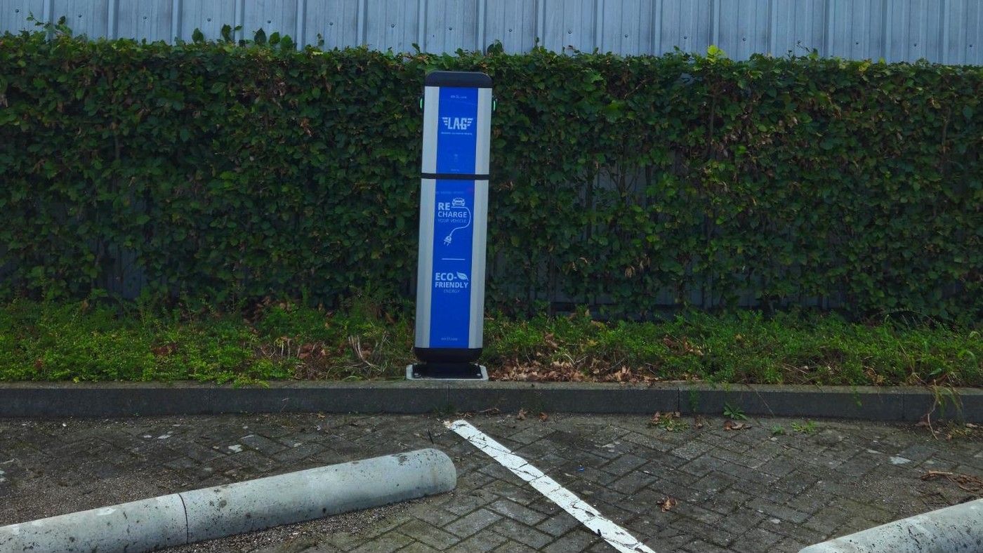 Laadpaal met blauw paneel voor een haag, met parkeerplaatsen op de voorgrond