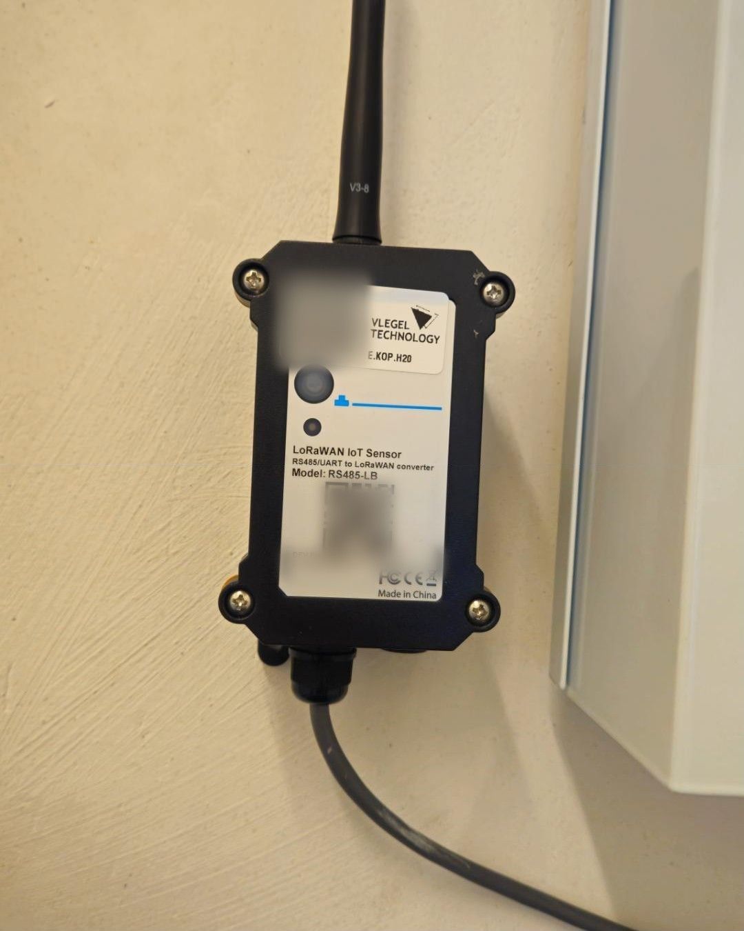Zwarte LoRaWAN-converter met antenne en label met meetpuntcode aan een wand