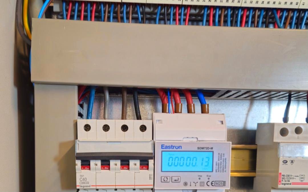 Energiemeter met verlicht display op DIN-rail naast een vierpolige automaat, onder een klemmenstrook