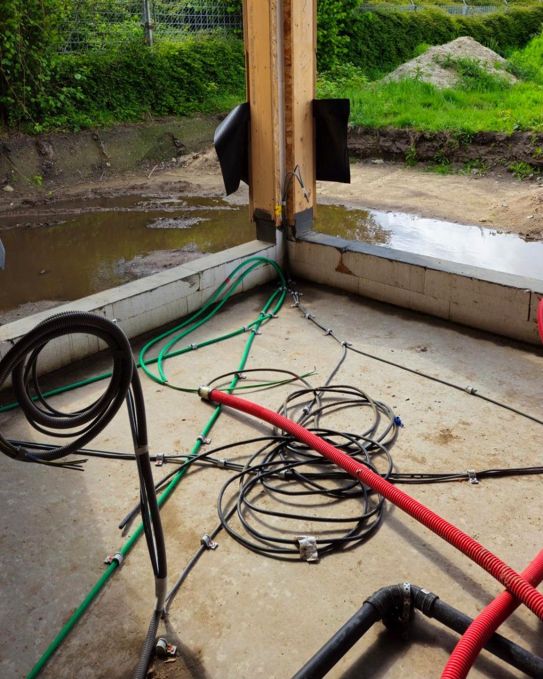 Hoek van een houtskeletwoning in opbouw met groene en zwarte kabels en rode buizen op de betonvloer