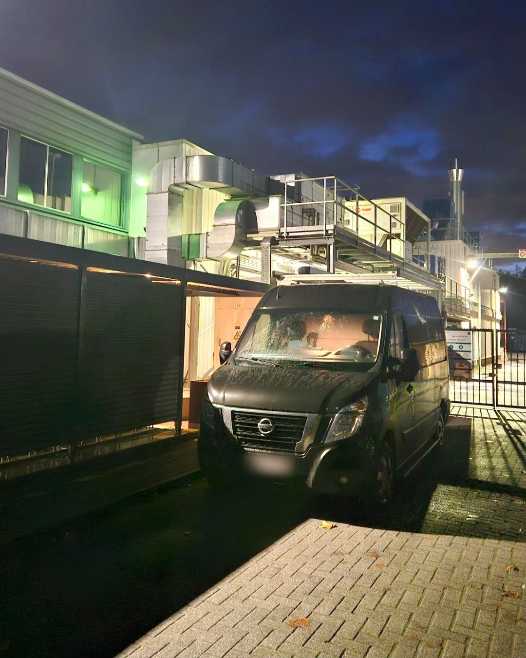 Donkere bestelwagen 's avonds op een bedrijfssite, voor een gebouw met verlichte technische installaties